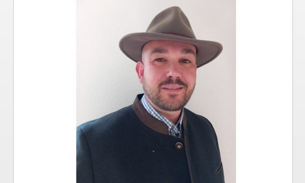 Ein Mann trägt einen Hut und eine traditionelle Jacke, er schaut in die Kamera, Hintergrund ist eine weiße Wand.