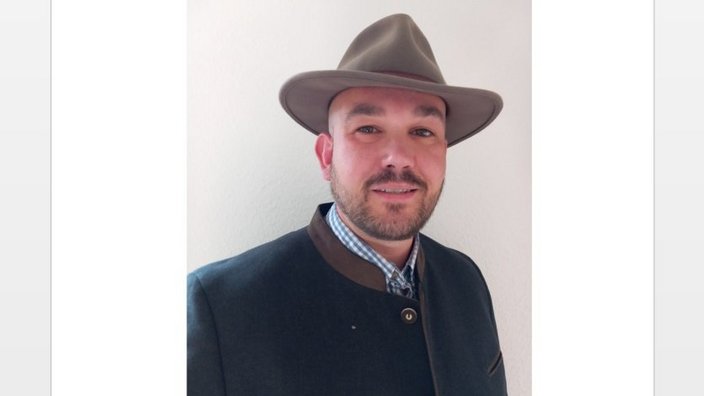 Ein Mann trägt einen Hut und eine traditionelle Jacke, er schaut in die Kamera, Hintergrund ist eine weiße Wand.