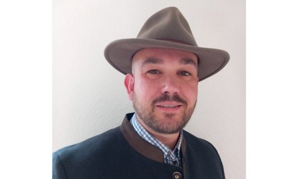 Ein Mann trägt einen Hut und eine traditionelle Jacke, er schaut in die Kamera, Hintergrund ist eine weiße Wand.